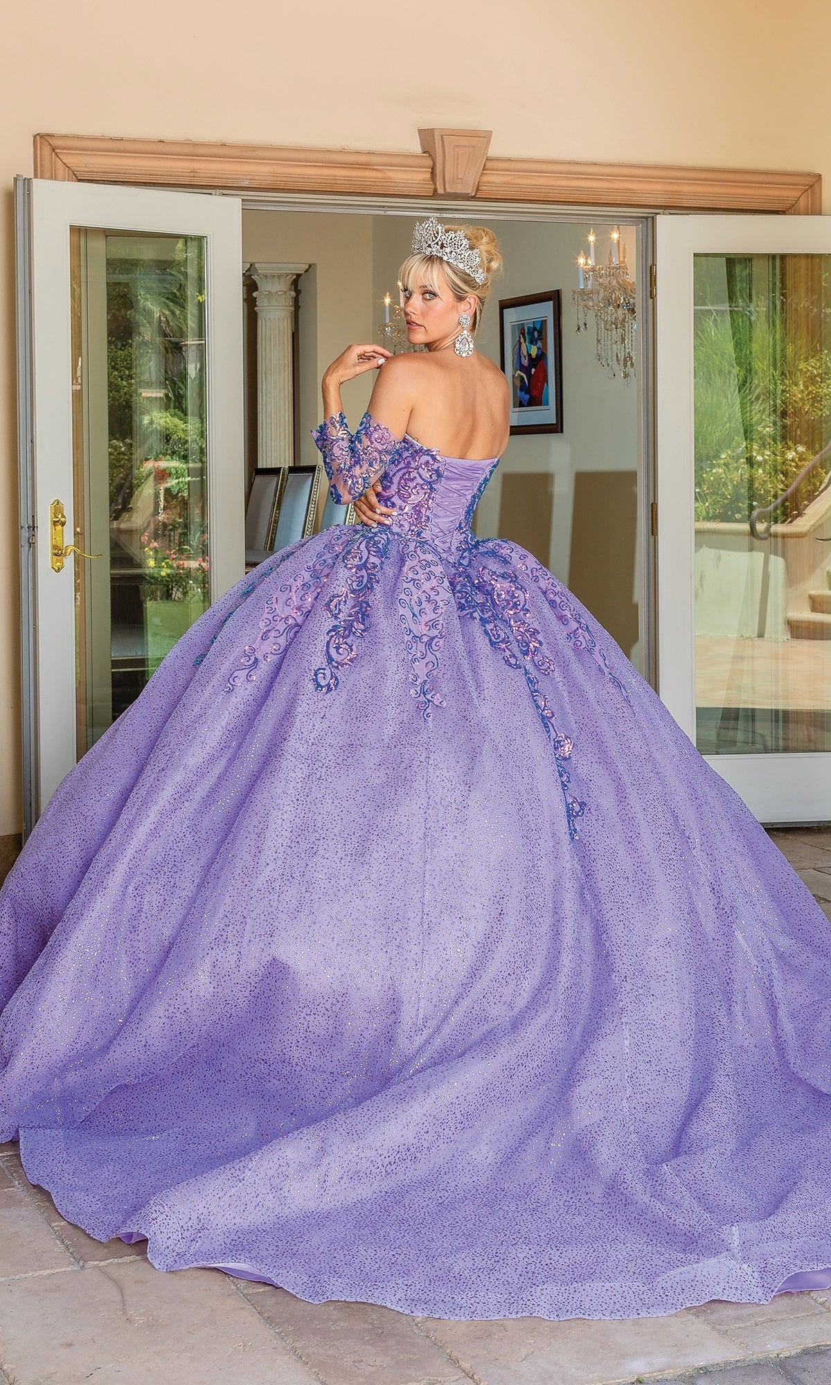 A woman wears the Quinceanera Dress 1675 By Dancing Queen, a large, ornate lavender ball gown with floral details and a tiara, standing indoors near open glass doors with her back to the camera.